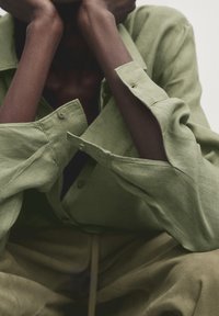 Personne à la peau foncée portant une chemise boutonnée oversize verte claire et un pantalon vert, reposant ses bras et sa tête sur ses mains.
