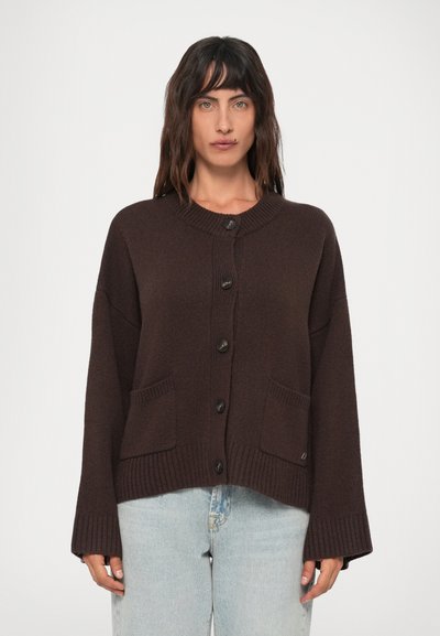 Modèle féminin portant un cardigan en tricot marron foncé à boutons avec deux poches avant et un jean bleu clair, se tenant contre un fond blanc.
