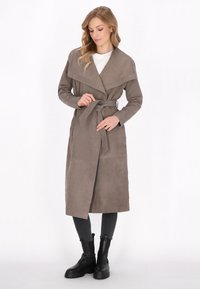 Lang beige frakke med en bred krave, bæltestrop i taljen og to sidelommer. Fremstillet af blødt stof med en glat tekstur. Båret over mørke jeans og støvler.