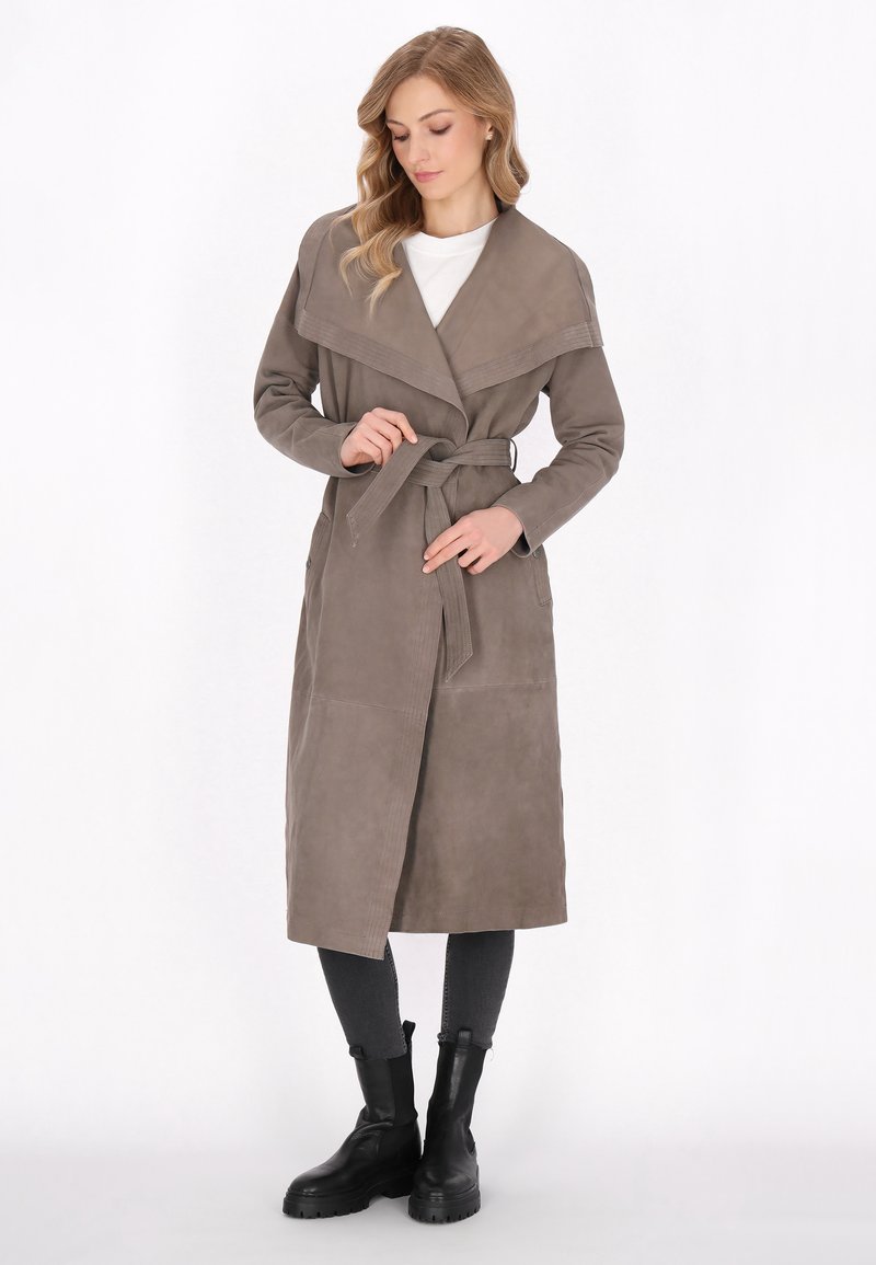 Lang beige frakke med en bred krave, bæltestrop i taljen og to sidelommer. Fremstillet af blødt stof med en glat tekstur. Båret over mørke jeans og støvler.