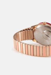 Montre en or rose avec un bracelet métallique segmenté comportant des maillons alternant entre finition mate et polie. Boîtier rond avec une lunette rouge et un fond métallique.