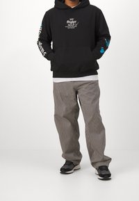 Černá mikina s kapucí s bílým nápisem na přední straně a modrým motivem motýla na rukávu, doplněná o šedé pruhované kalhoty a černé tenisky.