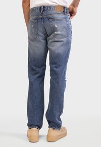 Jeans di denim blu con una vestibilità rilassata, caratterizzati da dettagli strappati, cinque tasche e una patch in pelle marrone sulla vita posteriore.