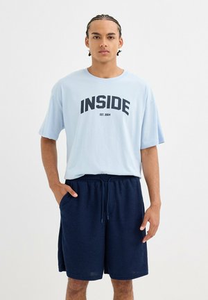 Joven con cabello trenzado de pie, vistiendo una camiseta azul claro de gran tamaño con el texto "INSIDE EST. 2004" y pantalones cortos azul marino con cordón.
