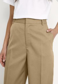 Bruna vida byxor i lätt tyg, med sidofickor och en slät textur, stiliserade med en vit topp.