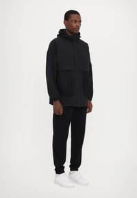 Svart zip-up jacka med huva, med nätfickor och en strukturerad design. Matchad med svarta joggers och vita sneakers.