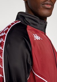 Czerwona i czarna kurtka z kołnierzem na zamek, z logo Kappa na piersi oraz wzorzystymi wstawkami po bokach, wykonana z gładkiego poliestru.