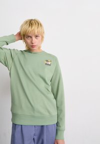 Sweatshirt vert clair avec un col rond, des poignets et un ourlet côtelés. Présente un petit motif imprimé de fleurs sur le côté supérieur gauche.