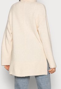 Krémová oversized svetr s vysokým límcem, ribbed spodním lemem a švem, který vedle probíhá vertikálně dolů přes záda. Měkká textura.