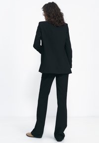 Kobieta z kręconymi włosami, ubrana w czarny dopasowany garnitur i beżowe buty, stoi i zwrócona jest tyłem do obiektywu, przodem do białej ściany.