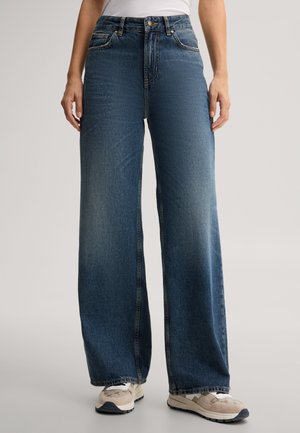 Persona che indossa jeans blu a vita alta e gamba larga con bottoni e cerniera davanti, abbinati a sneaker beige e bianche.