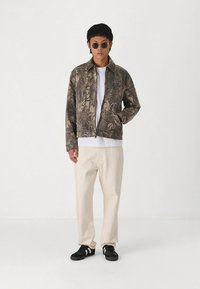 Veste à imprimé camouflage en tissu texturé, associée à un t-shirt blanc et un pantalon ample de couleur claire, complétant le look avec des baskets noires.