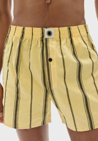 Gula randiga badshorts med elastisk midja, med svarta vertikala linjer och knappstängning. En sol-logotyp syns på midjan.