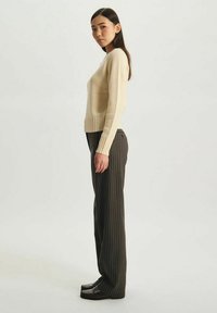 Pull en maille beige clair avec un ourlet côtelé, associé à un pantalon large rayé sombre et des chaussures noires. Le mannequin se tient de profil devant un fond uni.