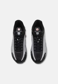 Nike Shox tenisice s gornjim dijelom od crne mrežice i srebrnim sintetičkim premazima. Imaju okrugle vezice, jastučiće na jeziku i prepoznatljiv dizajn potplata.