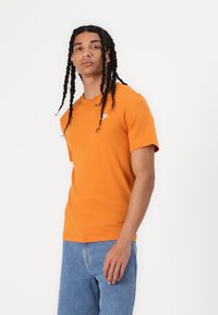 Oranžs kokvilnas T-krekls ar apaļu kakla izgriezumu un īsām piedurknēm. Uz kreisā krūts ir neliels balts logotips. Apvienots ar zilām džinsu biksēm.