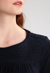 Mörkblå topp med en texturerad spetsdetalj i axeln som har blommönster, rund halsringning och rynkad tyg under axeldetaljen.