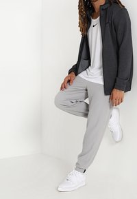 Pantalons de sport gris, veste zippée gris foncé et baskets blanches. Les matériaux semblent lisses avec une texture minimale. Logo Nike visible sur le t-shirt.