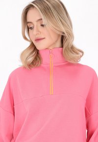 Rosa Reißverschluss-Sweatshirt mit hohem Kragen, ausgestattet mit einem kontrastierenden gelben Reißverschluss. Weicher Stoff mit lockerer Passform und ohne auffällige Muster.