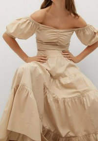 Beige off-the-shoulder mekko, jossa on puffihihat, kerätty rintaosa ja kerroksellinen röyhelöhelma, valmistettu pehmeästä kankaasta, jossa on hienovarainen tekstuuri.