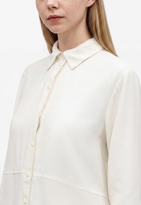 Femme à la peau claire et aux cheveux blonds portant un chemisier blanc à col boutonné, regardant vers l’avant avec une expression neutre.