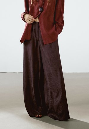Persoon draagt losse, donkerbruine wijde broek en een gestructureerde roodbruine blouse met knopen, waarbij één hand de zoom van de blouse aanpast.
