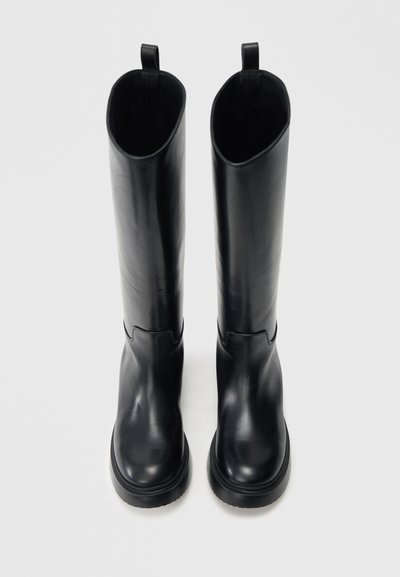 Μαύρες μπότες μέχρι το γόνατο από λείο δέρμα. Έχουν στρογγυλή μύτη, παχιά σόλα και λουράκι στην κορυφή για εύκολη τοποθέτηση.