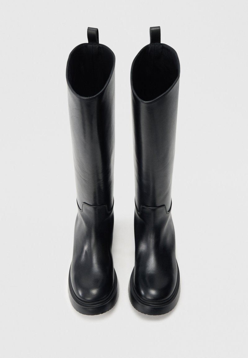 Stivali neri alti fino al ginocchio in pelle liscia. Presentano una punta arrotondata, suola spessa e una linguetta in cima per facilitarne la calzata.