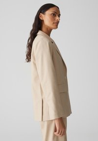 Beige op maat gemaakte blazer van gestructureerde stof. Heeft een reverskraag, twee voorzakken en een enkele knoopsluiting.