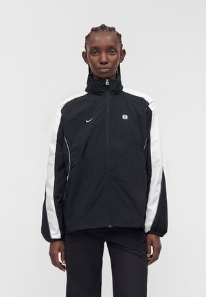 Kvinde med flettet hår iført en sort Nike trackjakke med hvide striber på ærmerne, stående mod en ensartet lys grå baggrund.