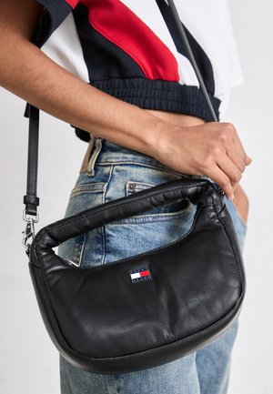 Mano sosteniendo un bolso de hombro de cuero negro de Tommy Jeans, combinado con vaqueros azules y un top corto en rojo, blanco y negro.