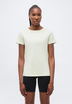 Femeie cu părul lung prins în coadă, purtând un tricou deschis la culoare cu mâneci scurte și pantaloni scurți negri pentru ciclism, stând în fața unui fundal simplu.