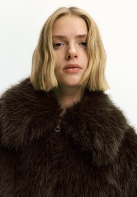 Mladá žena s krátkými blond vlasy, oblečená v tmavě hnědém koženém kabátu, s neutrálním výrazem, na jednoduchém světlém pozadí.