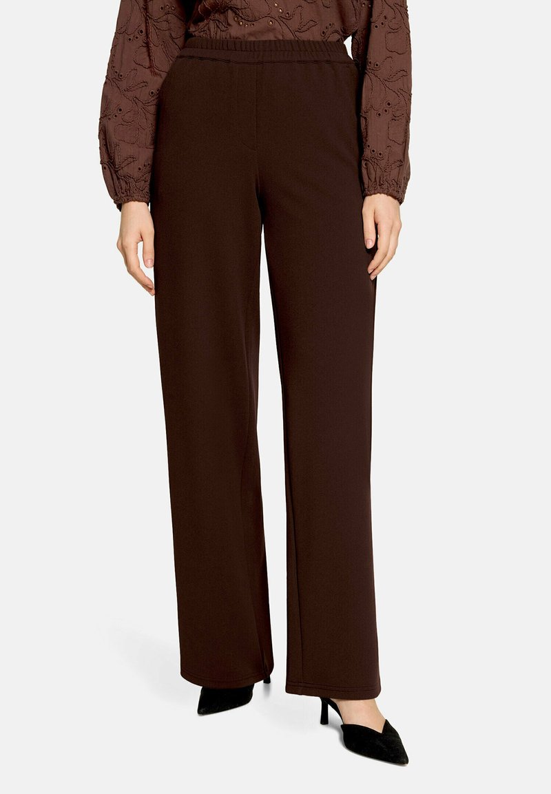 Femme portant un pantalon ample marron foncé avec une taille élastique, un chemisier marron à motifs à manches longues, et des talons pointus noirs.