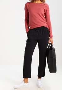 Top de manga larga rosa claro con hombros de encaje texturizado, combinado con pantalones negros de cintura elástica y zapatillas blancas, sosteniendo un bolso negro.