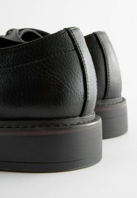 Zapatos de cuero negro con superficie texturizada, tacones redondeados y detalles de costura. Las suelas están ligeramente elevadas con un acabado suave.