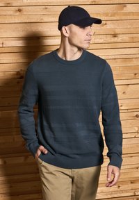 Dark blue textured sweater with horizontal ribbed patterns, crew neckline, and long sleeves, paired with light khaki pants and a cap.