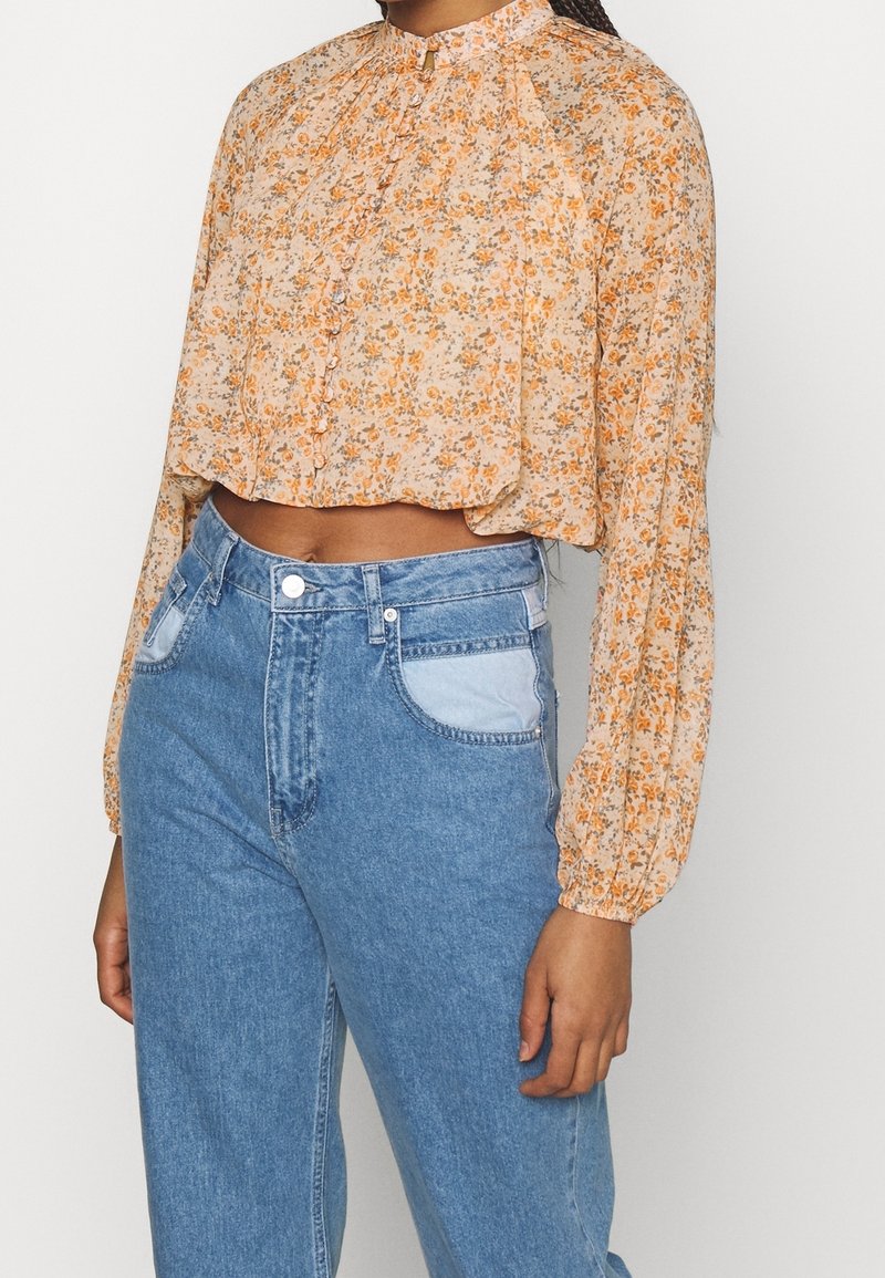 Blusa corta floral en naranja claro y crema, con largas mangas abullonadas, cierre de botones y delicados volantes en el escote.