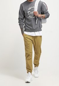 Felpa grigia con grafiche bianche, pantaloni tapered beige e sneakers bianche. L'outfit presenta una vestibilità comoda ed è completato da uno zaino.