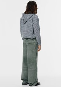 Lichtblauwe geruite hoodie met capuchon, gecombineerd met losse, vervaagde groene broek. Gestructureerde stof en twee achterzakken zichtbaar. Zwarte schoenen.