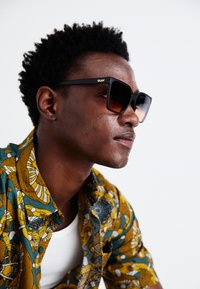 Black oversized sunglasses with gradient brown lenses. The subject wears a patterned yellow and green shirt, featuring abstract designs and textures.