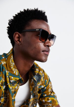 Schwarze oversized Sonnenbrille mit braunen Farbverlauf-Gläsern. Das Subjekt trägt ein gemustertes Hemd in Gelb und Grün, das abstrakte Designs und Texturen aufweist.