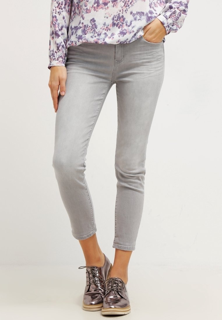Jeans ajustados de color gris claro con una textura suave, que presentan una cintura de altura media, una silueta entallada y un corte hasta los tobillos, combinados con zapatos brillantes.