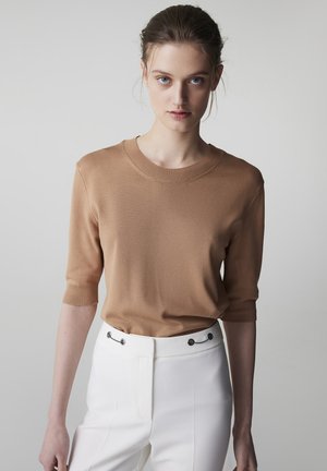 Jeune femme aux cheveux attachés portant un pull à manches courtes couleur chameau et un pantalon blanc taille haute sur un fond clair.