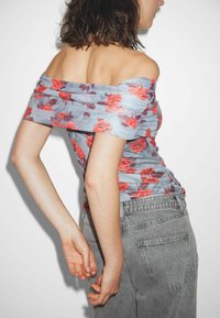 Donna in top floreale off-shoulder con rose rosse e jeans grigi a vita alta, in posa di lato su uno sfondo bianco semplice.