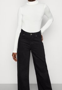 Vit långärmad turtleneck-top som kombineras med högmidjade, vida svarta jeans med fem fickor och metalldetaljer.