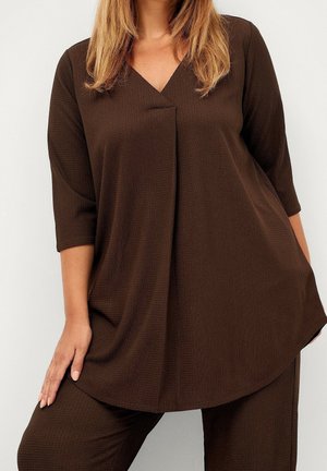 Žena má na sobě volné tmavě hnědé texturované tričko s tříčtvrtečními rukávy a odpovídající kalhoty, ruka spočívá na stehně.