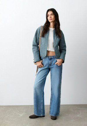 Mujer de pie con chaqueta acolchada azul, top corto blanco, vaqueros anchos azules, zapatos marrones y bolso beige al hombro, frente a una pared lisa.