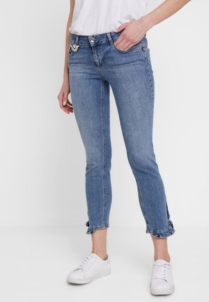 Kvinde iført blå skinny jeans med flæsekant, en hvid t-shirt, hvide sneakers og en dekorativ broche på brystlommen.