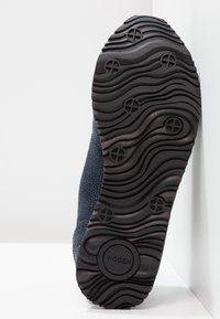 Sneaker z teksturowaną granatową cholewką i czarną gumową podeszwą z falistym wzorem oraz wytłoczonym logo w okrągłym akcentem.